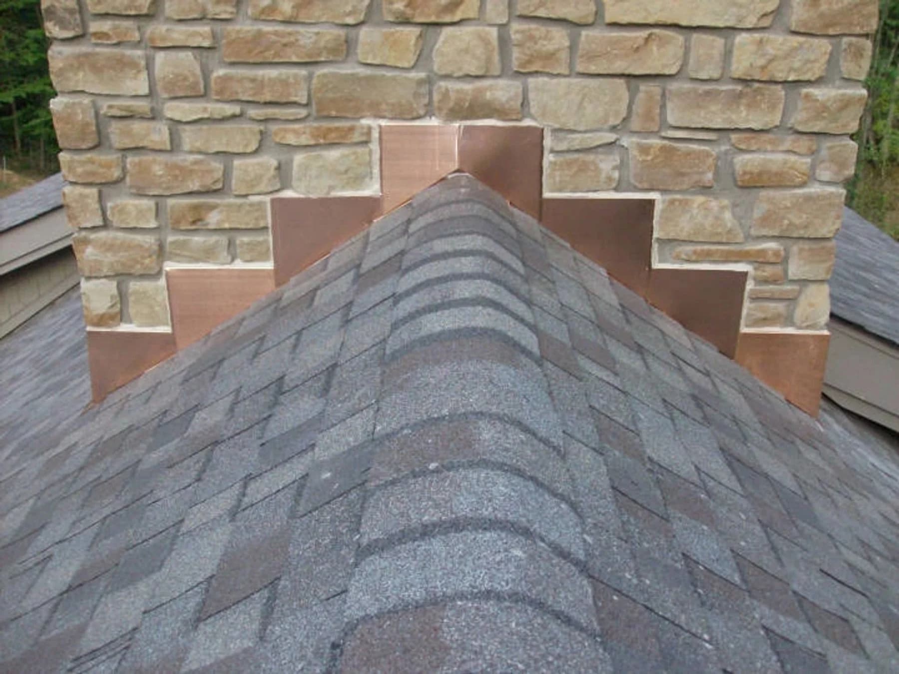 Tan stone chimney with stepped copper-colored flashing and a shingled roof cricket