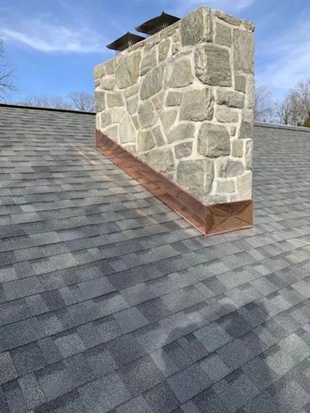 Gray fieldstone chimney with copper-colored flashing where it meets the shingle roof